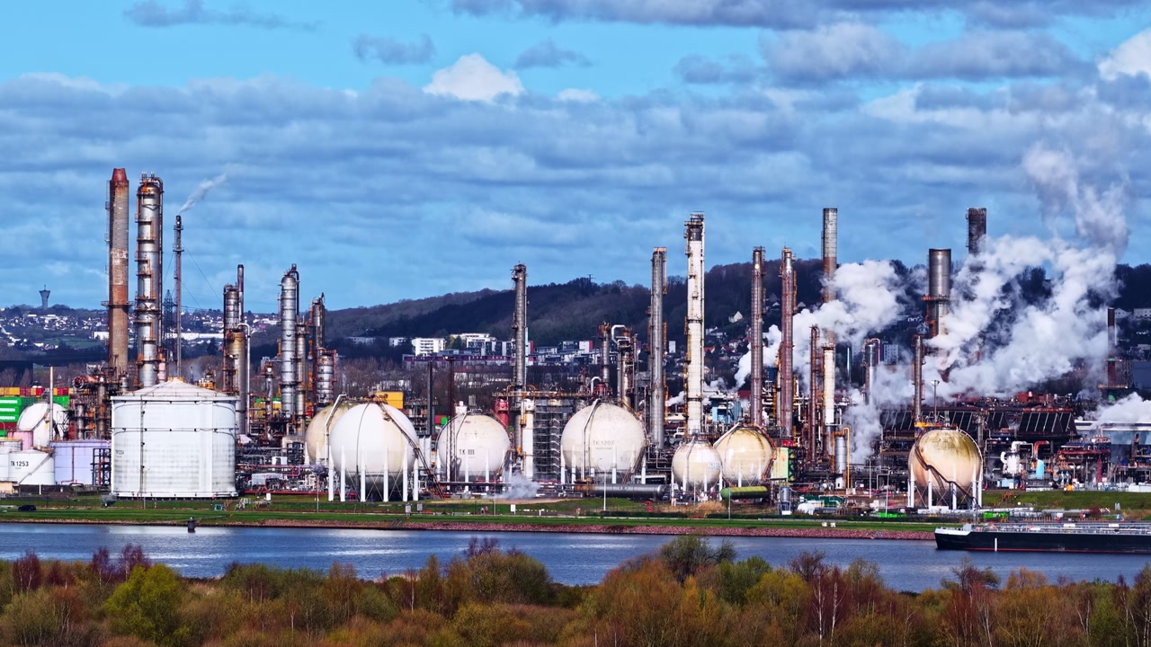 Petrochemical refinery with storage tanks towers and steam emissions along industrial waterfront, aerial, static time lapse