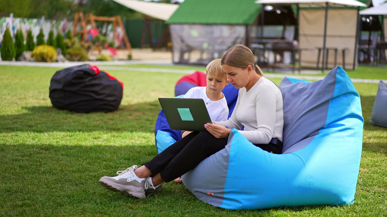 Mother and her child sitting in the green lawn in comfortable chairs. People looking at the computer screen attentively.