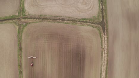 Vista-Aérea-Hacia-Abajo-Desde-Un-Dron-Que-Se-Cierne-Sobre-Un-Campo-Agrícola-Marrón-Mientras-El-Tractor-Remolca-Un-Equipo-A-Través-Del-Suelo-Debajo