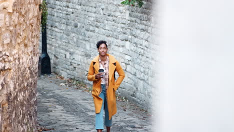 Front-view-of-young-black-woman-wearing-a-yellow-pea-coat-holding-a-takeaway-coffee-walking-towards-camera-in-an-alleyway-between-stone-walls,-elevated-view