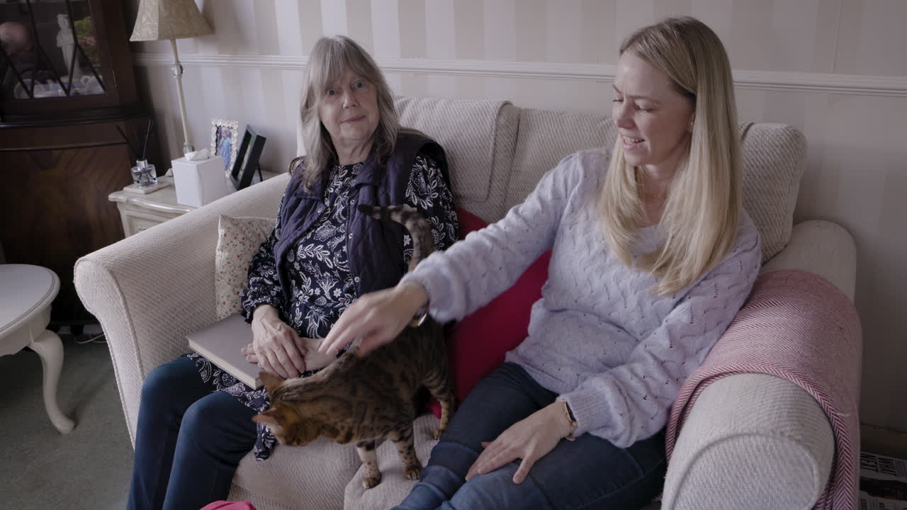 Two women sitting on a couch petting a cat