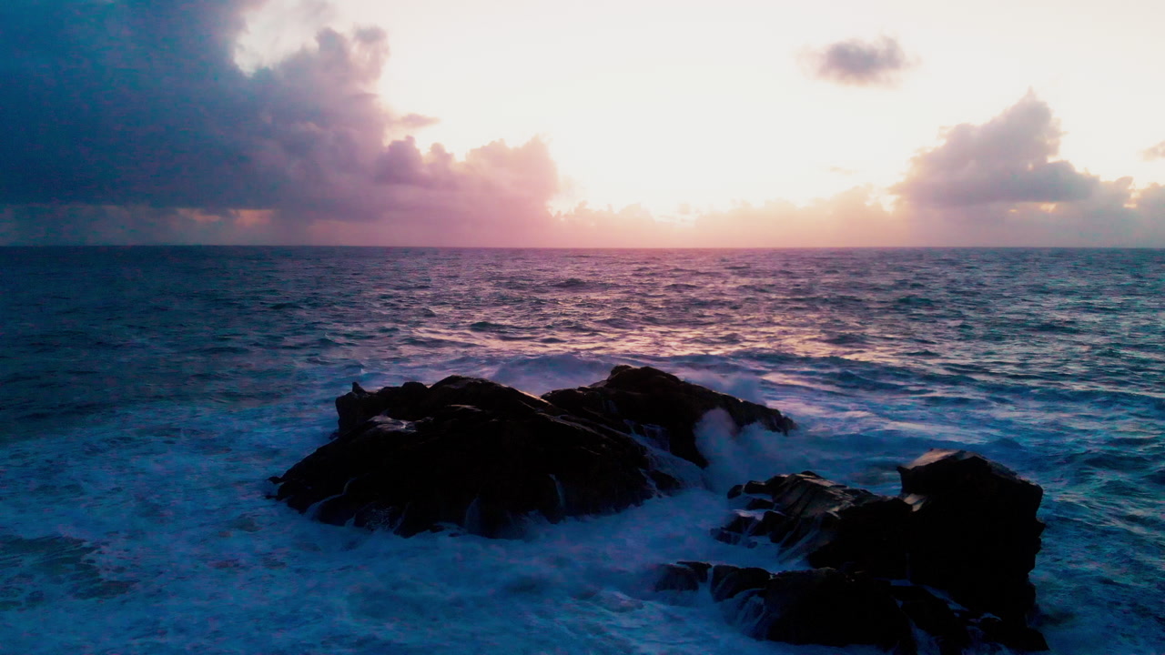 Low angle drone shot near water level showing waves crashing on rocks with sunrise over ocean horizon