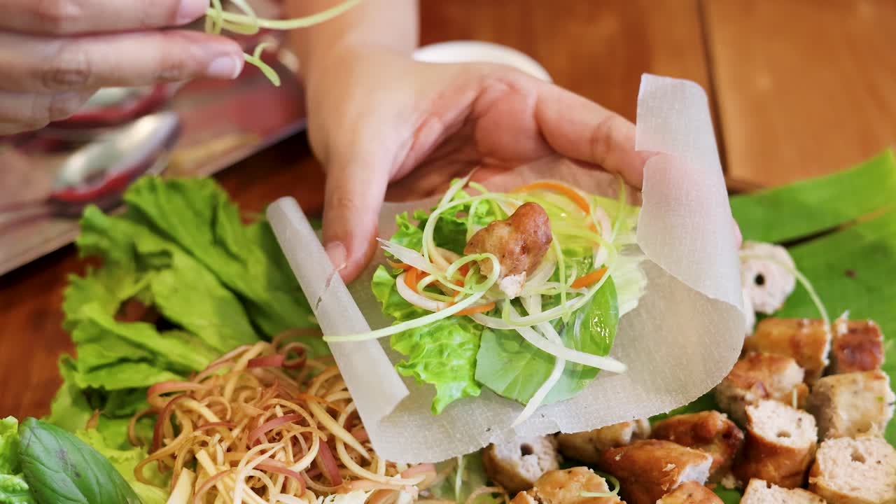 Bidikan jarak dekat tangan yang dengan hati-hati membungkus bahan-bahan segar ke dalam kertas nasi untuk masakan tradisional Vietnam