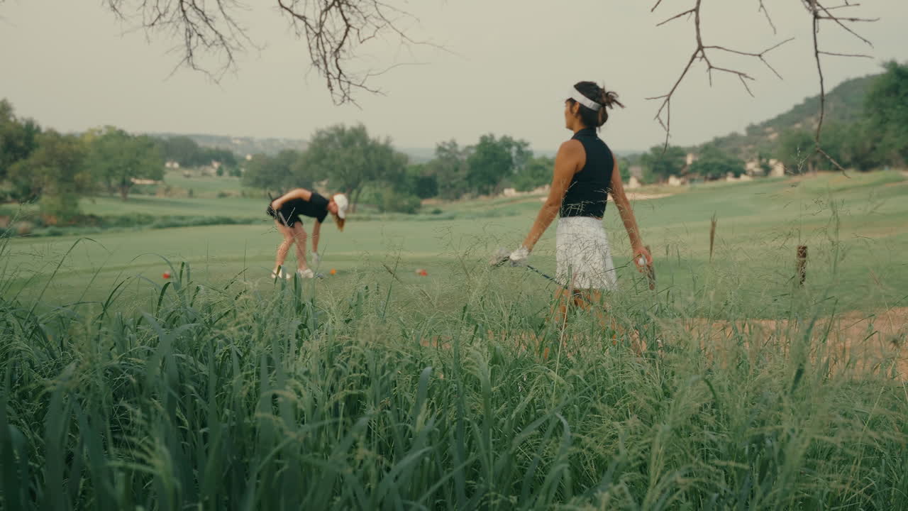 Two lady golfers on the tee box preparing to start their round. One places her tee in the ground while her female partner walks up. Outdoor golf course scene on a sunny day in slow motion