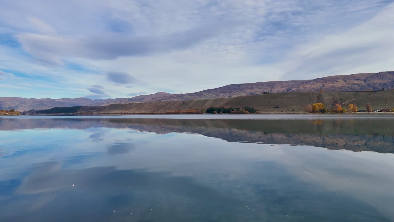 Aerial footage captures Lake Dunstan's tranquil waters and autumn foliage under a dynamic sky in Cromwell, New Zealand