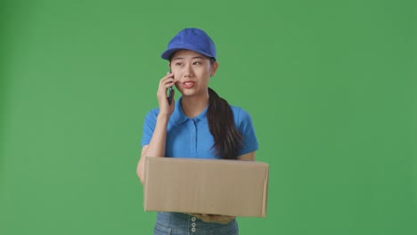 asian female courier in blue uniform talking on smartphone while delivering a carton on green screen background in the studio