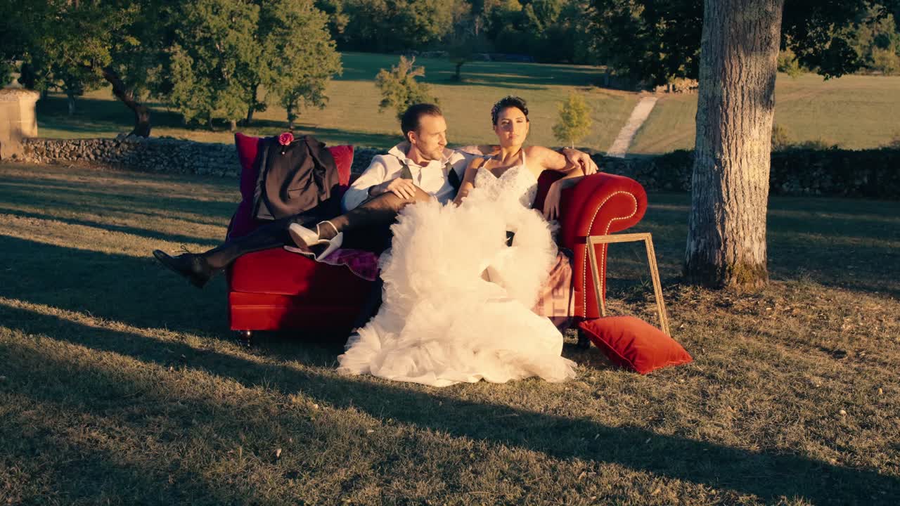 Nygift par sidder sammen på en rød sofa placeret midt på en grøn mark i naturligt lys på deres bryllupsdag. Totalbillede.