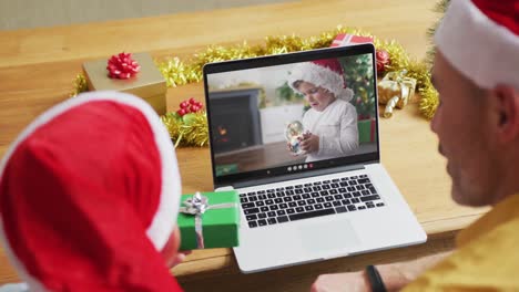 Padre-Caucásico-Con-Hijo-Con-Gorros-De-Papá-Noel-Usando-Una-Computadora-Portátil-Para-Una-Videollamada-Navideña,-Con-Un-Niño-En-La-Pantalla