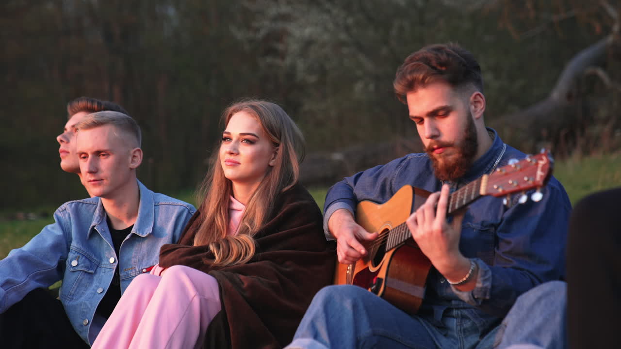 One bearded young man among his friends playing guitar. Other guys listening to music and enjoying it. Nature backdrop.