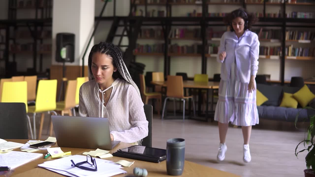 Open public library. Happy brunette in white casual clothes in headphones funny dancing on background while her friend concentrated on her studying, sitting at the table works with laptop. Slow motion