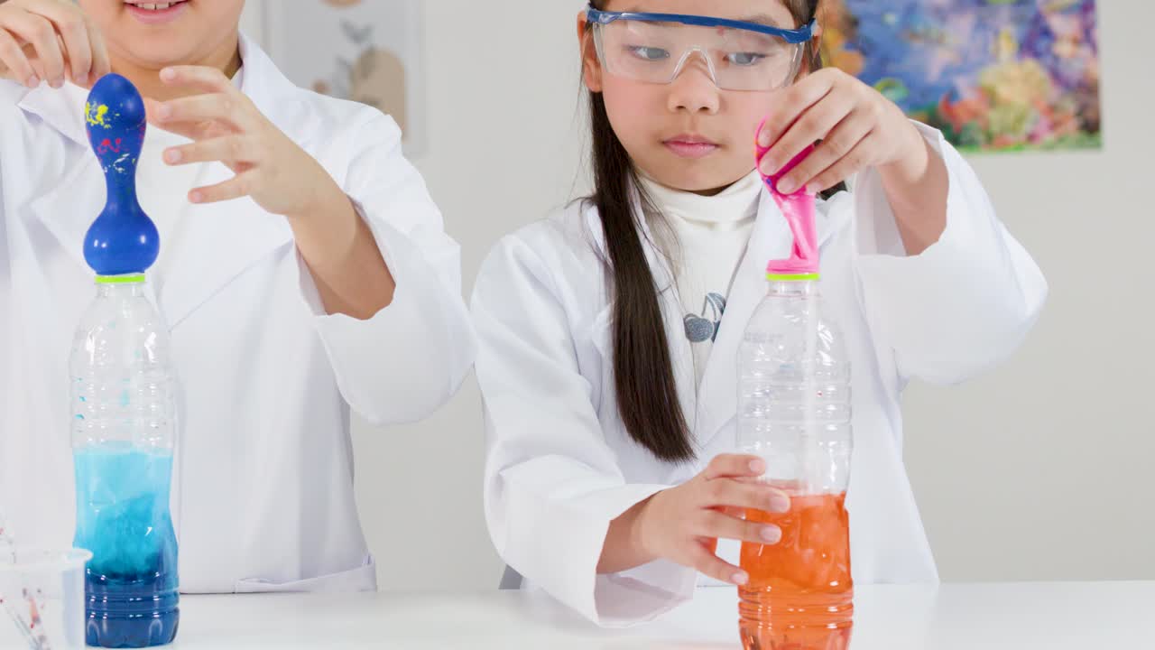 Two children in lab coats conduct a chemical reaction experiment inflating balloons on plastic bottles