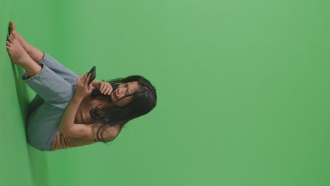 full body of young asian woman victim of violence with bruise on body looking at smartphone and crying while sitting in the green screen background studio