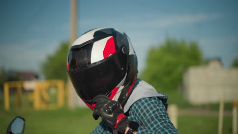 una motociclista femenina con casco y guantes de protección negros baja su visera, preparándose para montar, lleva equipo completo, con un fondo borroso