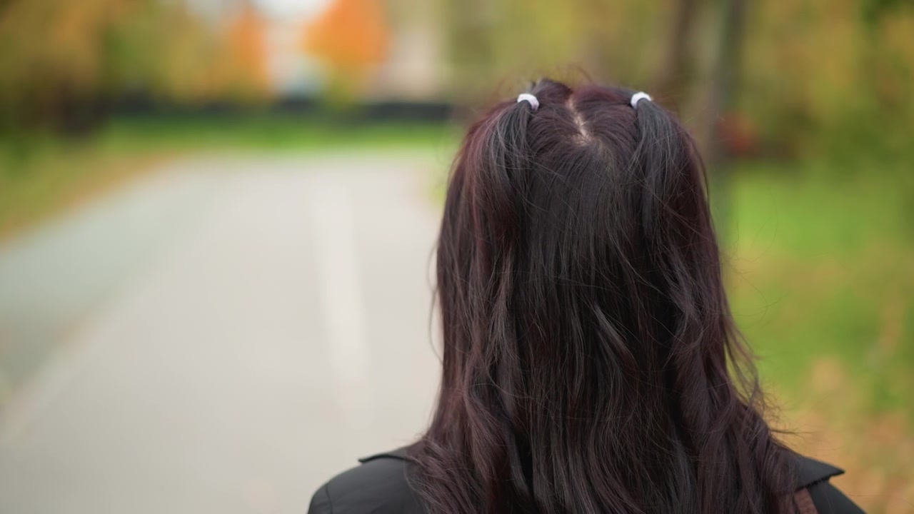 Nærbillede af en ung pige med langt mørkt hår stylet i to fletninger, der går væk på en udendørs sti. Hendes ryg og hoved er i primært fokus, hvilket viser naturlig bevægelse i blødt lys.