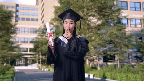 asian woman student graduates in cap and gown holding diploma and making shh gesture in front of a magnificent university building