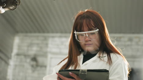 lab technician wearing face shield observes lifted car while taking notes on laptop in mechanical workshop, background features industrial tools, mechanical equipment, and structured workspace