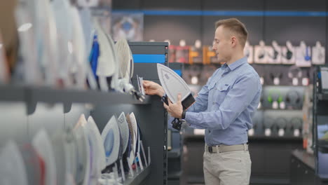 A-young-handsome-man-in-a-shirt-chooses-an-iron-in-a-consumer-electronics-store.-Bachelor