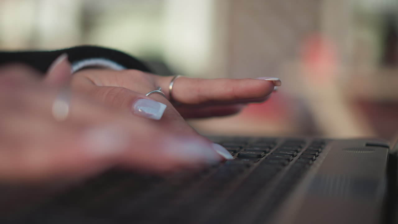 Close-up of hands typing on a laptop keyboard with blurred background, focusing on fingers and nails, the view shows the action of typing with polished nails and delicate rings