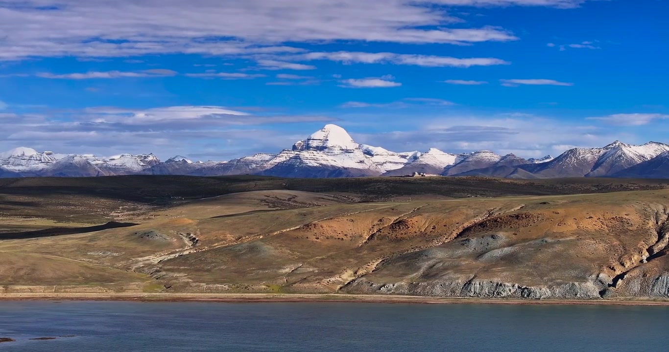 Et lateralt drone-glideskud fanger det levende blå vand i Manasarovar-søen langs Kailash-bjergkæden i Tibet, der afslører tørt terræn, bølgende bakker og sneklædte tinder