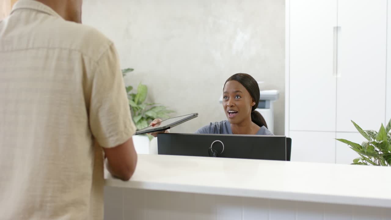 Receptionist afroamericana accoglie un visitatore che si avvicina da sinistra mentre controlla il tablet per la verifica dell'identità.
