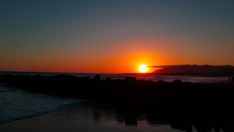 Slow-and-low-shot-rising-above-rocks-and-revealing-sunset-with-silhouettes-of-people-by-the-shore-at-San-Buenaventura-State-Beach-in-Ventura,-California,-United-States