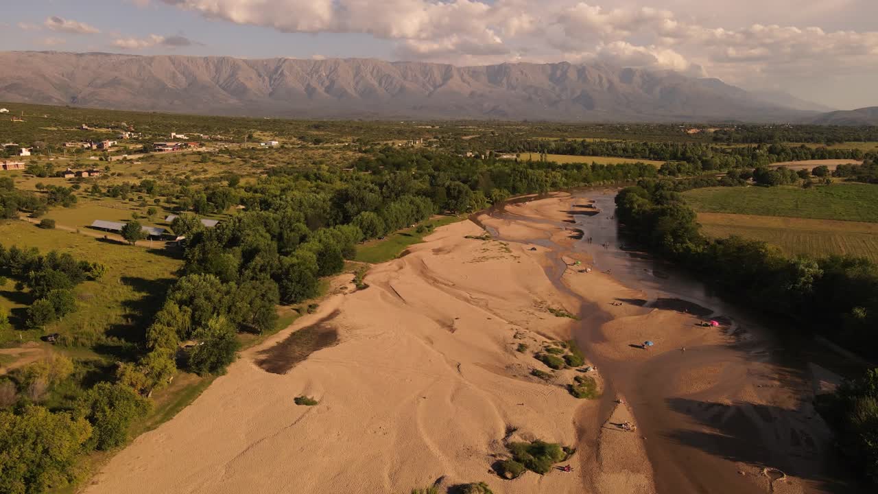 Pemandangan 4K 60 fps: Sungai Berpasir dengan Tepian Hijau dan Pegunungan di Latar Belakang saat Matahari Terbenam