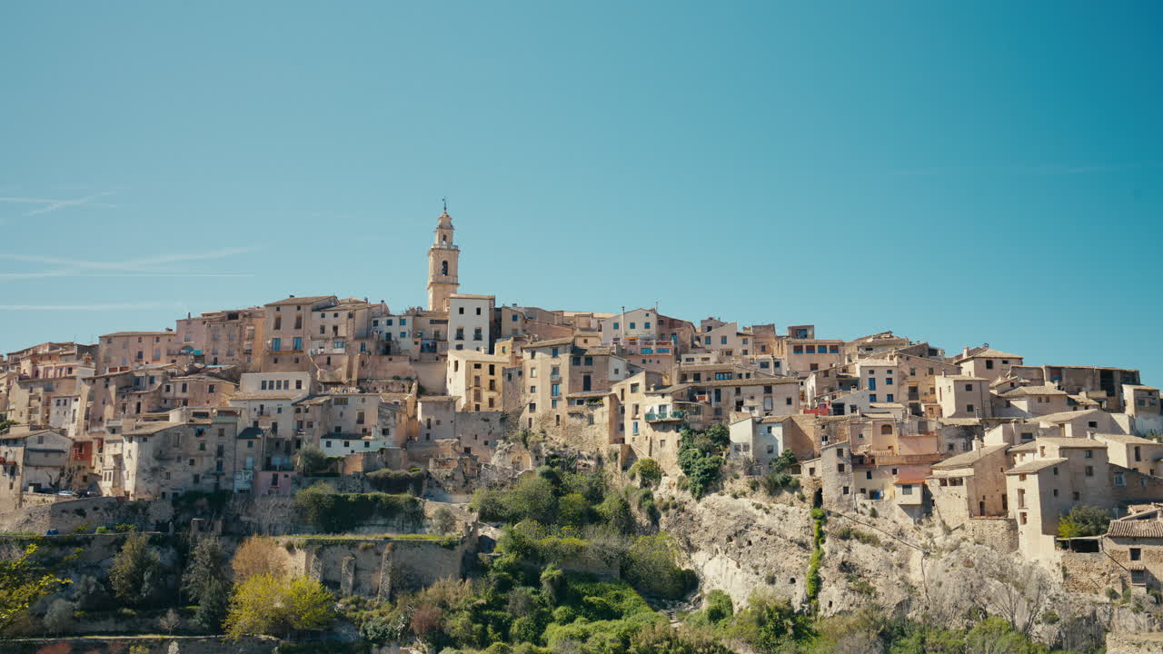 Una vasta vista panoramica mostra Bocairent, Spagna, che si estende sulla collina rocciosa sotto un cielo azzurro e limpido, con case a strati e un campanile che definiscono lo skyline.