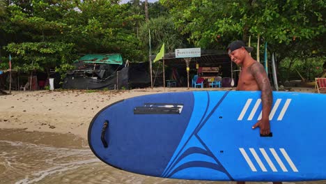 walking-tracking-shot-of-happy-Asian-man-walking-on-the-beach-with-a-stand-up-paddle-board-on-a-beach-with-watersports-shop-in-background