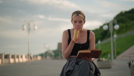 mujer sentada al aire libre con las piernas cruzadas sosteniendo un libro en una mano mientras come una hamburguesa y lee, el fondo presenta un poste de lámpara con un ambiente al aire libre sereno y borroso