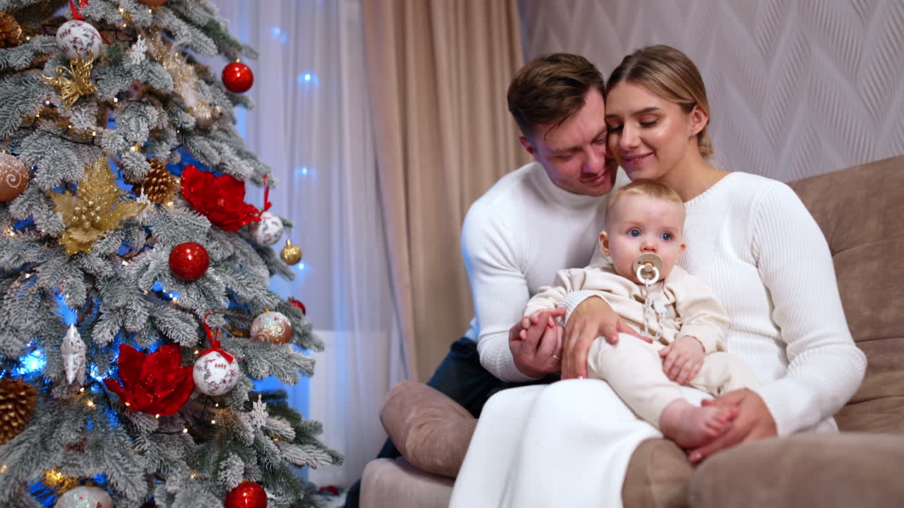 Happy family of three celebrating Christmas holidays together at home. Mom kisses her baby on the head while man kisses his wife.