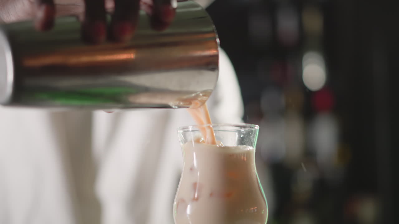 Bartender pouring drink from shaker into glass, creating a cocktail in a vibrant bar setting. Behind, illuminated bar shelves with liquor bottles and Heineken neon sign in the background