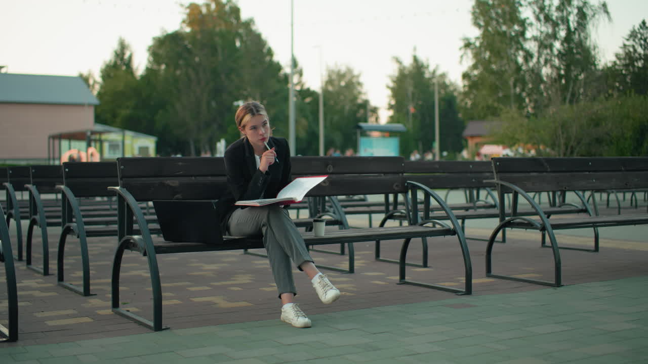 Young lady seated alone on outdoor bench deeply focused reading large open book while holding pen with laptop and coffee cup by her side in quiet urban setting surrounded by empty benches