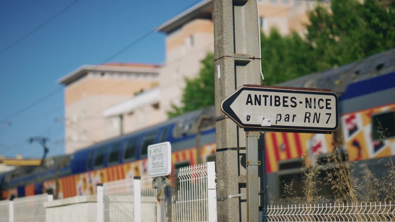 Primo piano di un cartello stradale Antibes-Nizza vicino ai binari ferroviari, con un treno colorato che passa sullo sfondo, catturando dettagli di trasporto e viaggio della Costa Azzurra.
