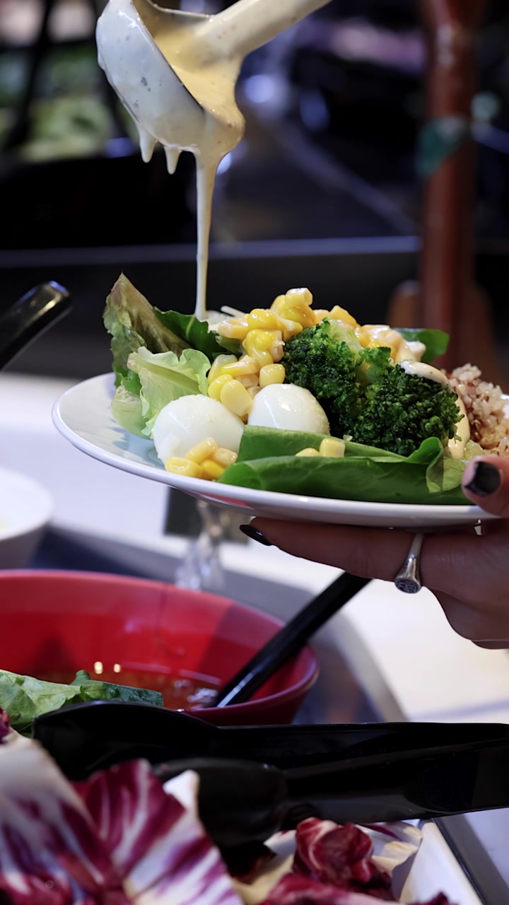 Primo piano di una mano che versa condimento cremoso su un piatto colorato di insalata da buffet, con messa a fuoco ridotta.