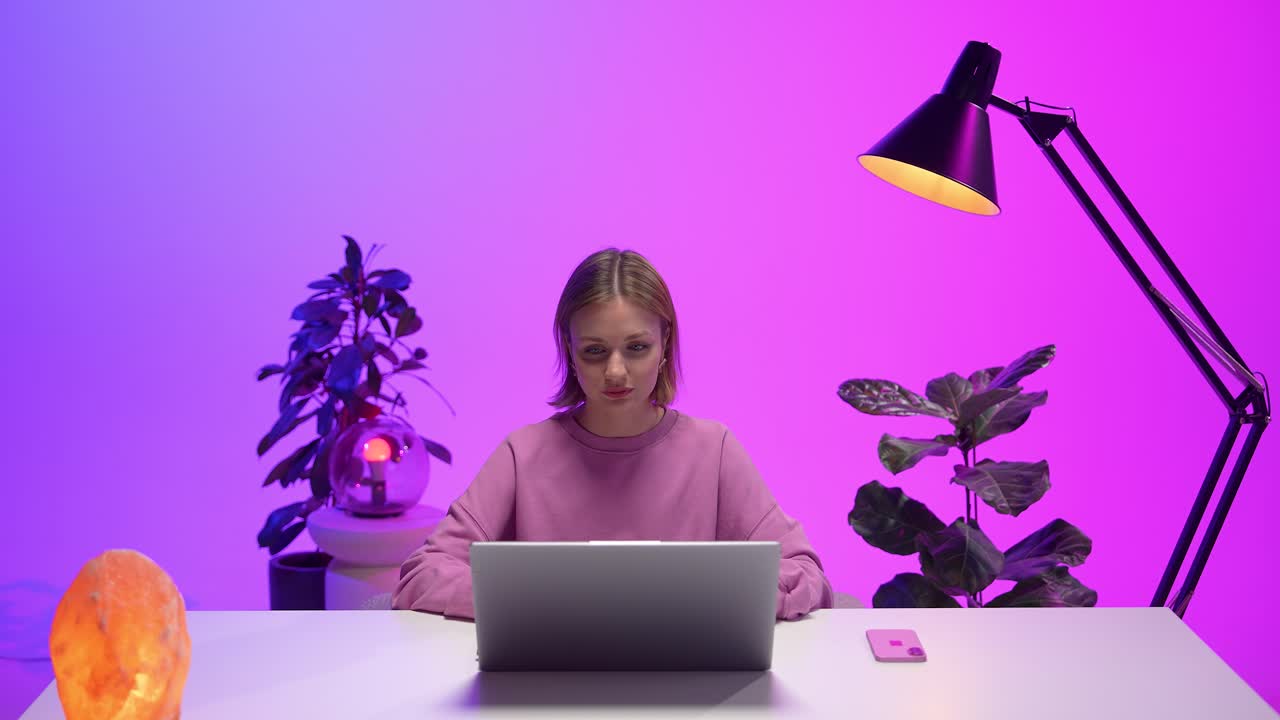 A woman sits at a minimalist desk working on a laptop, surrounded by glowing lamps and plants in a modern neon-lit interior. The ambiance blends tech focus with relaxing decorative elements