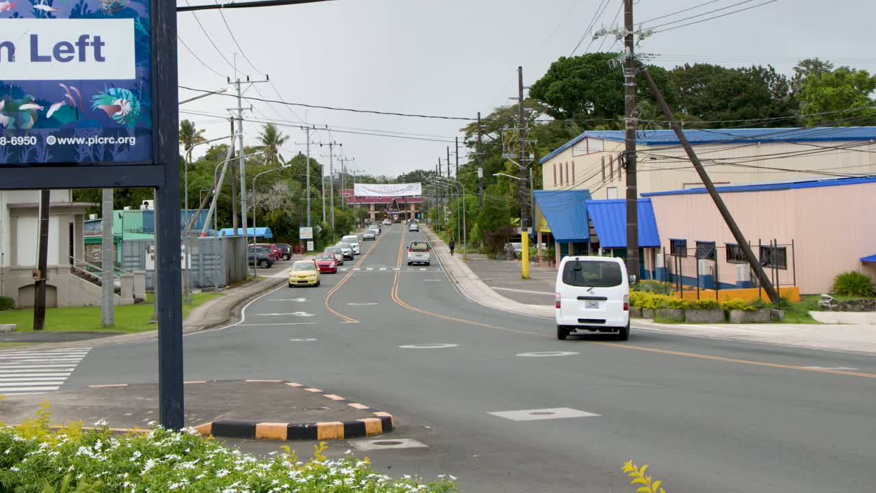 Một cảnh quay rộng tĩnh ghi lại những chiếc ô tô đang lái dọc theo con đường đô thị nhiệt đới dưới bầu trời nhiều mây.