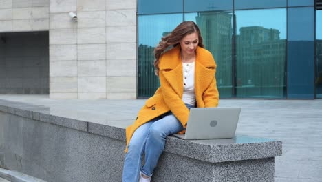 mujer de negocios usando portátil vestida con abrigo amarillo sentada al aire libre fondo de edificio corporativo
