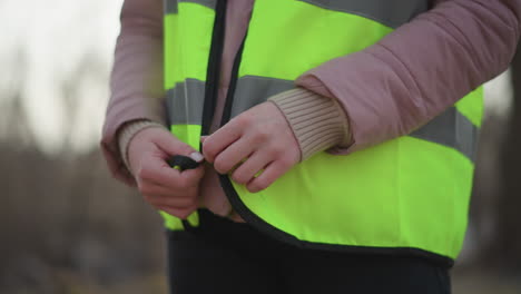 close-up of woman in pink winter jacket zipping bright yellow reflective safety vest outdoors in sunlight, preparing for work or volunteer task requiring visibility and protective clothing in park