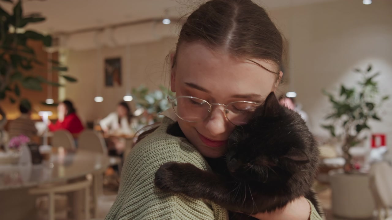 Primo piano di una donna che tiene in braccio un gatto nero in un accogliente caffè con illuminazione soffusa e avventori sfocati. La donna indossa un maglione di maglia verde e culla delicatamente il suo rilassato compagno felino tra tavoli, sedie e piante.