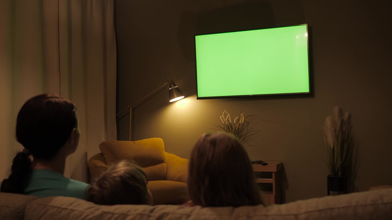Family Sitting Together Sofa In Their Living Room Night Watching TV Green Screen. Rear View Of Family With Children Sitting On Sofa In Living Room Evening Watching Green Mock-up Screen TV Together.