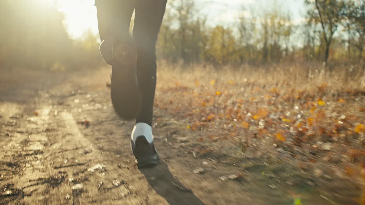 cerca de un hombre en un uniforme deportivo negro y zapatillas negras corre a lo largo de un camino de tierra en el bosque de otoño entre hojas marrones caídas y hierba seca en el otoño