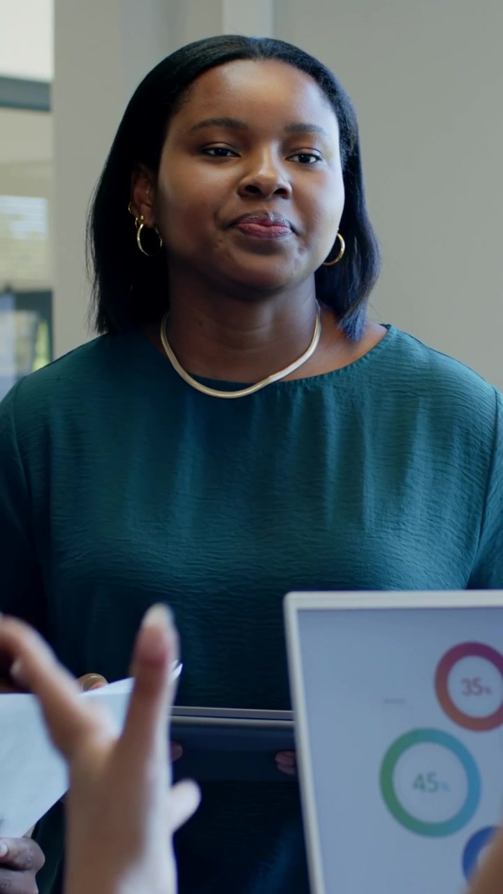 Vertical video: Noting teal-top woman holding docs and tab at desk reacting to peer point at charts
