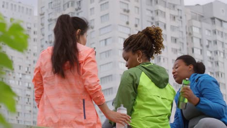 Foto-Trasera-De-Tres-Amigas-Multiétnicas-Con-Brillantes-Chaquetas-Deportivas-De-Neón-Sentadas-En-Un-Parapeto-Pedregoso,-Con-Un-Edificio-De-Apartamentos-De-Gran-Altura-En-El-Fondo,-Bebiendo-Agua-Y-Tomándose-Selfies-En-Un-Smartphone