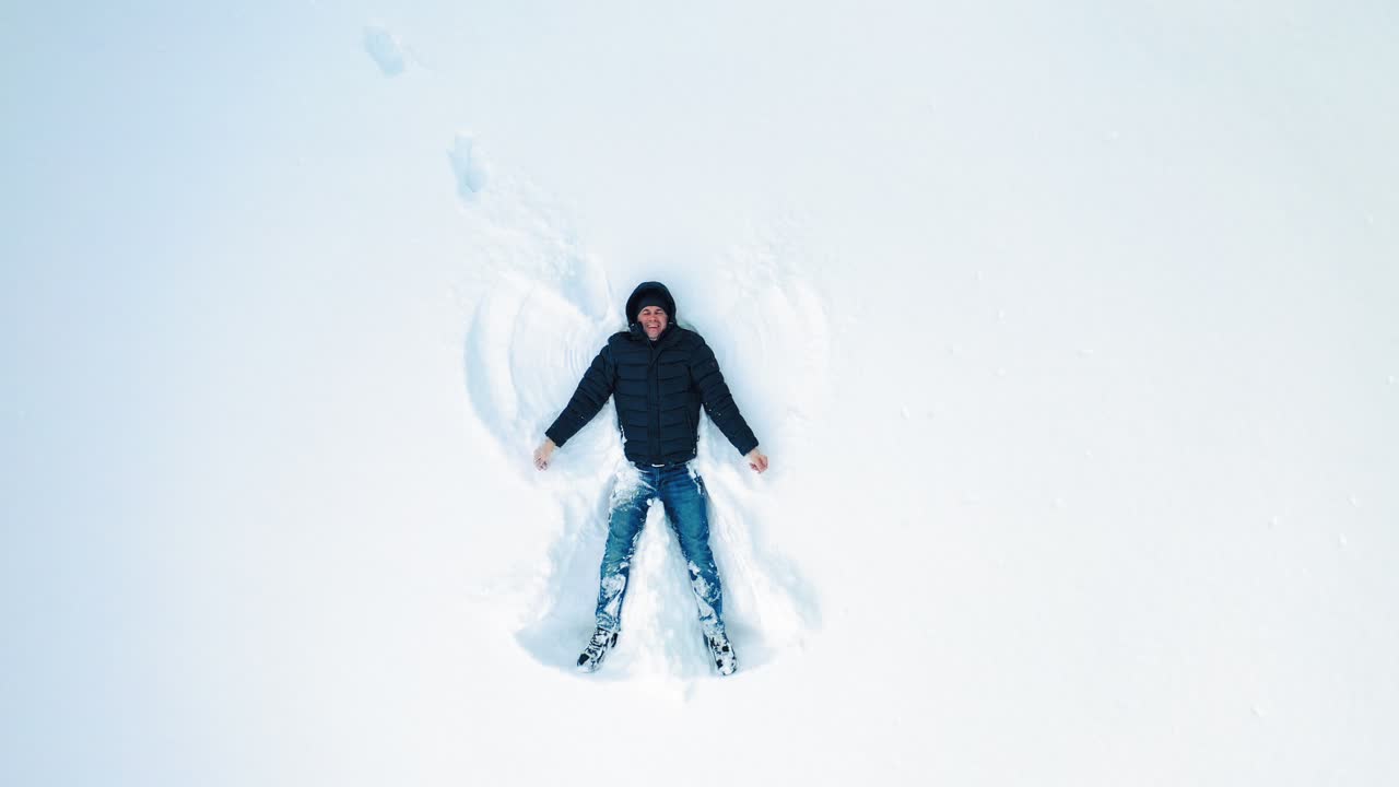 Adult man making snow angels. High angle view of happy man lying on snow and creating snow angel figure