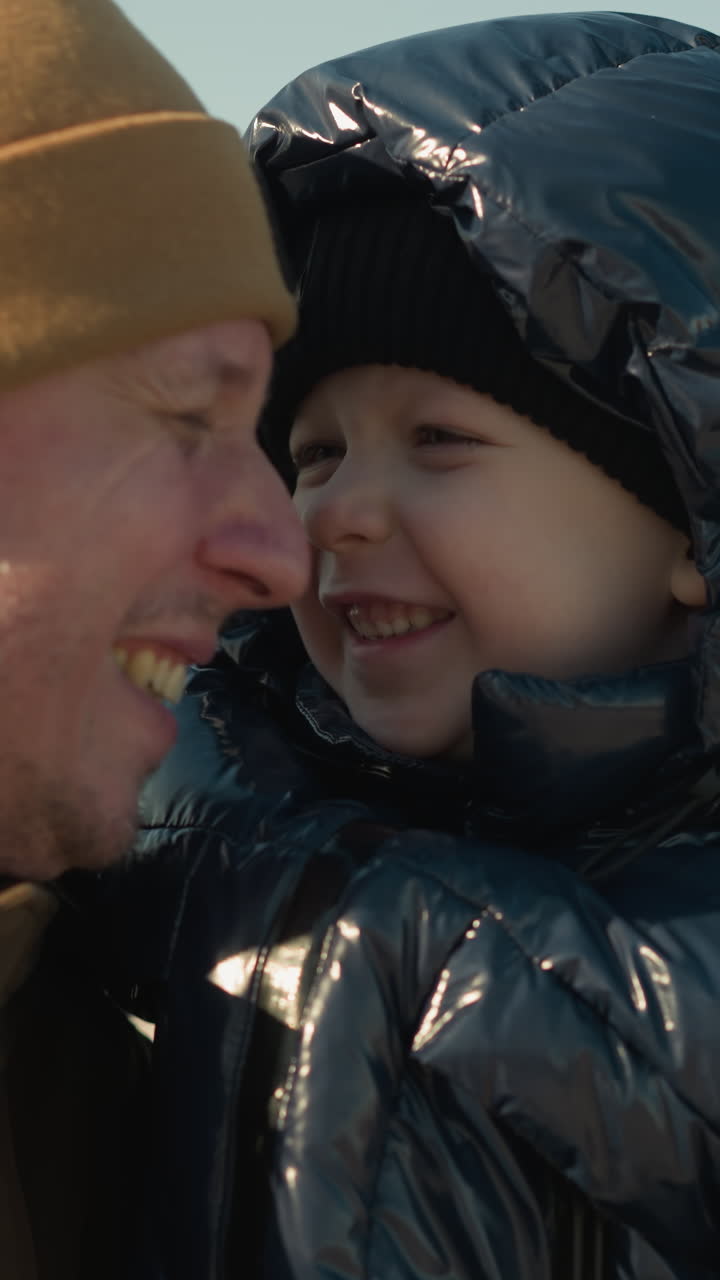 A joyful father carries his playful son close to his ear, with both of them smiling warmly, the child wears a black jacket, while the father wears a brown jacket and a beanie