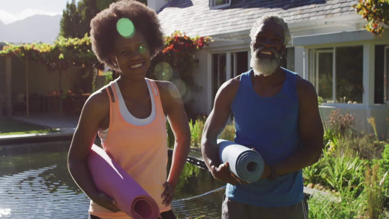 African american senior couple exercising outdoors carrying yoga mats in sunny garden