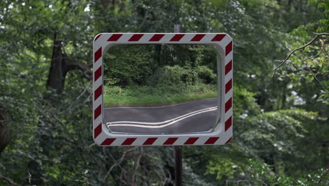 A-convex-road-safety-mirror-set-in-woodland-at-a-road-junction-as-vehicles-pass-by,-Worcestershire,-England