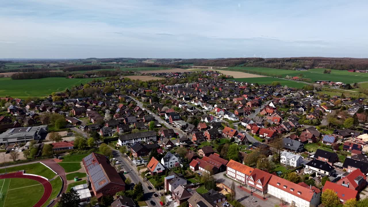 Weite Drohnen-Luftaufnahme einer deutschen Kleinstadt mit Wohnhäusern, einem Fußballfeld und der umliegenden Landschaft mit grünen Feldern und sanften Hügeln unter sanftem Frühlingslicht in einer ruhigen Vorstadtumgebung.