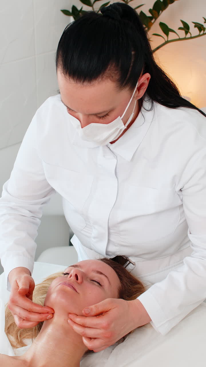 Cosmetóloga con mascarilla da un masaje facial en la línea de la mandíbula a una joven en un relajante spa de clínica de cosmetología