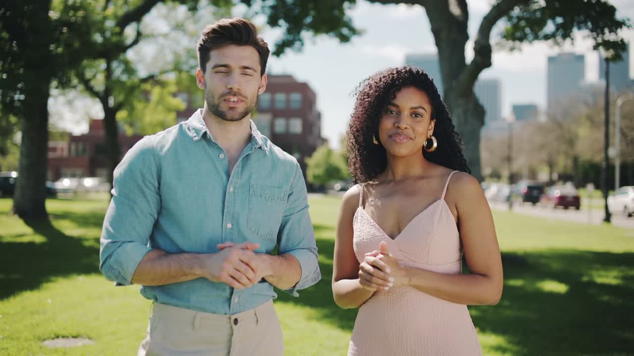 Câmera iniciando filmagem de homem de camisa azul e mulher de vestido rosa apresentando-se em um parque, mostrando o horizonte da cidade.
